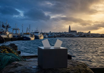 Black box with two white papers sits on a harbor pier, with fishing boats and a church across the water