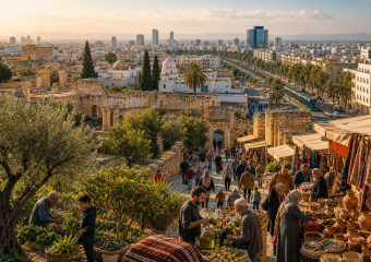 Busy market beside ancient ruins, with shoppers, pottery, textiles, olive trees, and a modern city skyline beyond