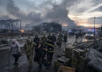 Firefighters escort civilians through the muddy ruins of a burning industrial site as smoke rises and emergency vehicles wait nearby