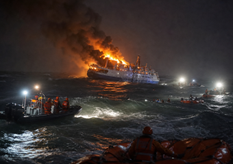 A passenger ship burns at sea as rescue boats approach through rough waves