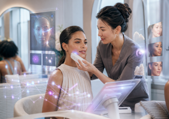 Aesthetician uses a handheld device to examine a woman's facial skin, with digital skin-analysis displays surrounding them