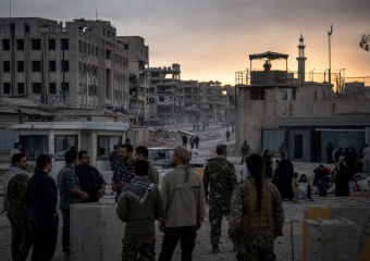 Civilians and armed personnel gather at a checkpoint amid war-damaged buildings at sunset