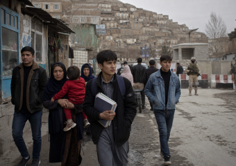 Civilians walk past armed soldiers on a city street below densely packed hillside buildings
