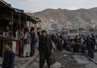 Crowded street market with shoppers, buses, and a mountainous cityscape in the background