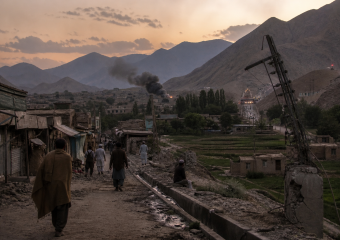 People walk along a rough road through a mountain valley settlement as smoke rises near fields at sunset