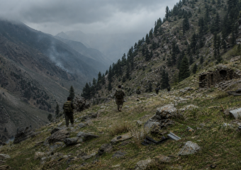 Soldiers hike along a rocky mountain trail beneath a cloudy sky
