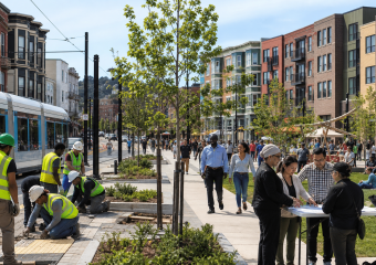 Workers plant trees beside a streetcar line as pedestrians walk through a lively urban neighborhood