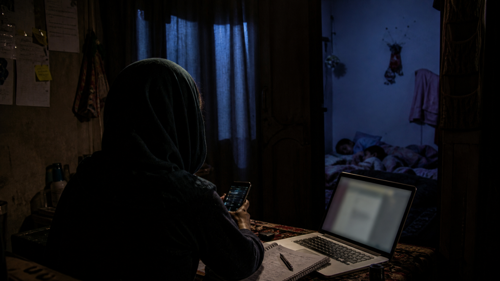 A person in a headscarf uses a laptop and phone at a table while two children sleep in an adjoining room