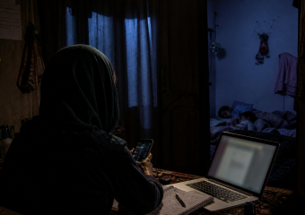 A person in a headscarf uses a laptop and phone at a table while two children sleep in an adjoining room
