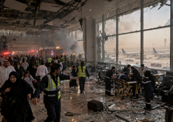 Firefighters and medics respond amid smoke and shattered glass in a damaged airport terminal, with planes visible outside