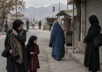Women and a child walk past a market street and security checkpoint with mountains in the background